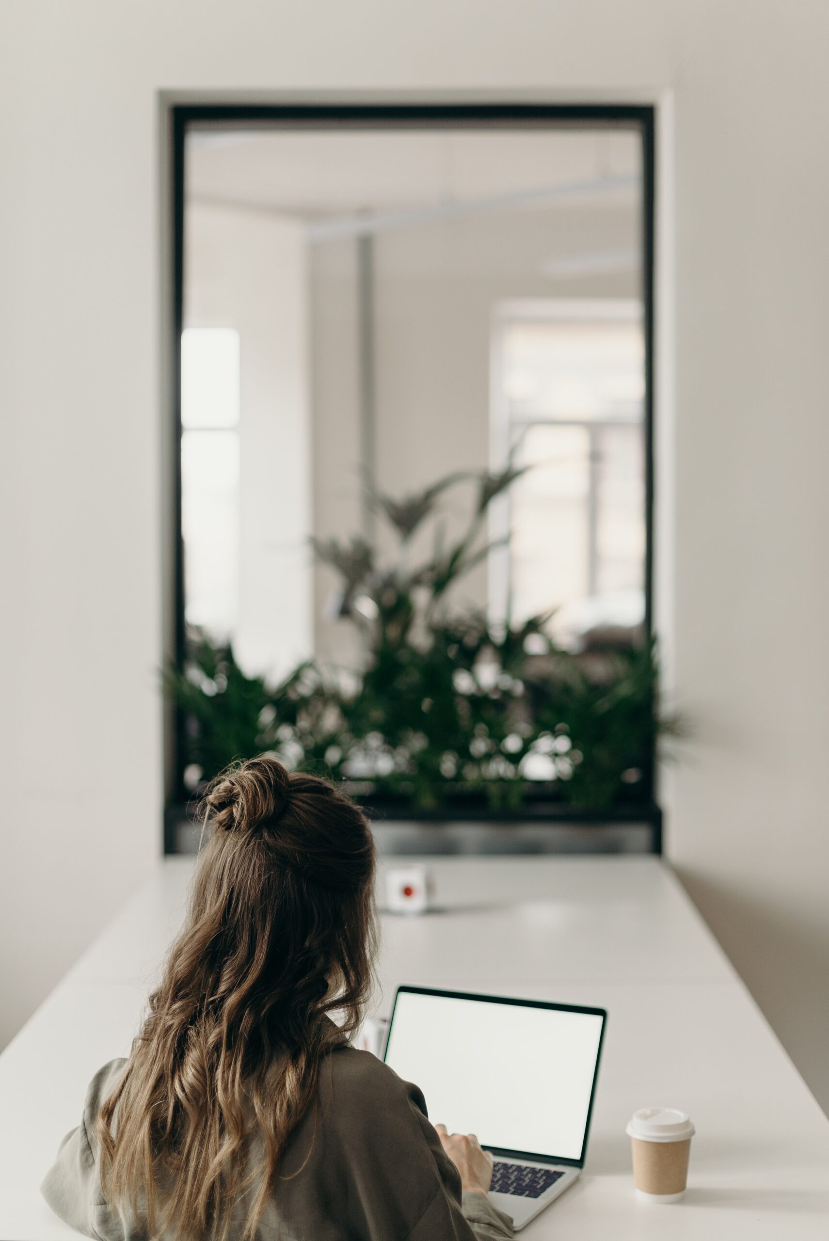 Woman sitting at table with laptop and coffee. Mirror and plants in background.