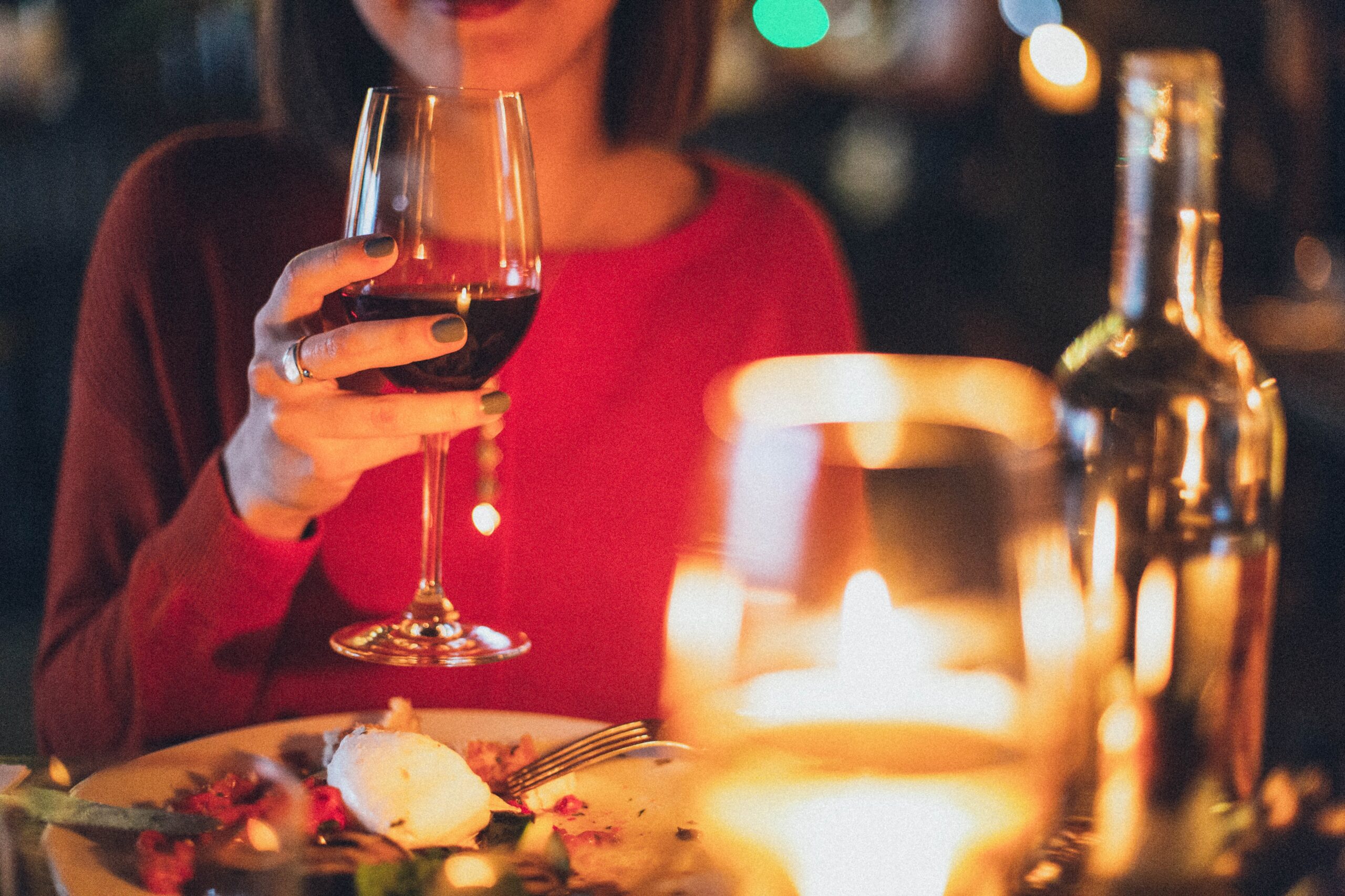 Close up of woman's hand holding a wine glass and dinner plate, with wine bottle and another glass out of focus in the foreground.