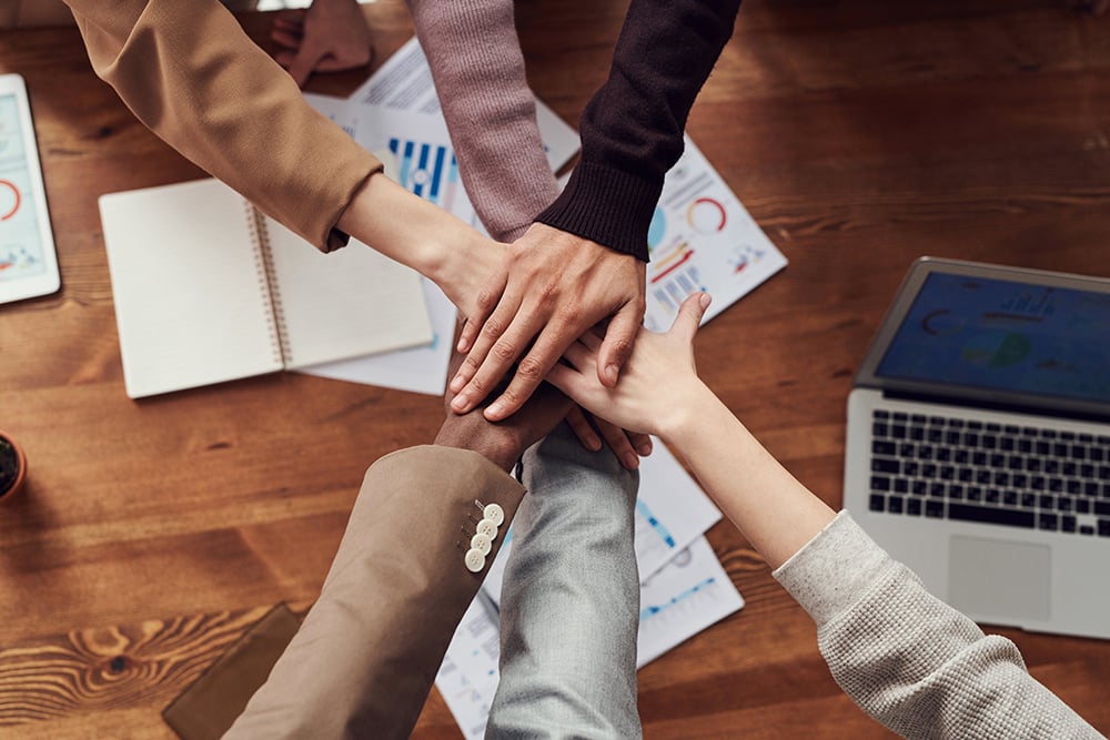 6 people stacking their hands to represent teamwork over a desk with papers and a laptop.