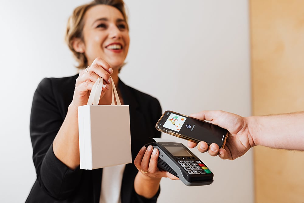 A woman hands a gift bag to a customer who is tapping their phone for payment on a handheld point of sale system.