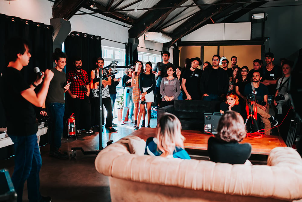 People standing, gathered around a person speaking in a large room.