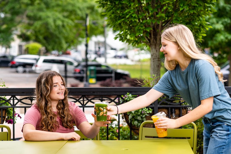 A young girl hands another a colorful drink.