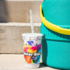 A stadium cup with a Color Run logo next to a portable water cooler on concrete steps.