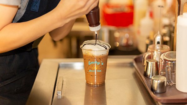 A barista adds chocolate sauce to the whipped cream, which tops an iced coffee in a custom printed plastic cold cup.