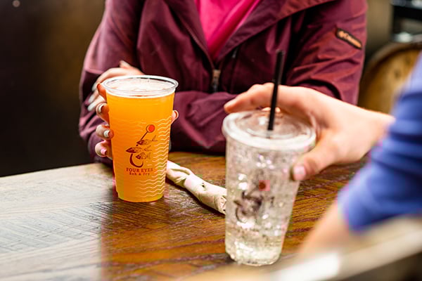 Two individuals sit at a table holding drinks, one with a beer, and another with a water.